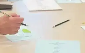 Na  długim stole leżą kolorowe zapisane kartki, oł&oacute;wki oraz dłonieuczestnik&oacute;w zajęć.