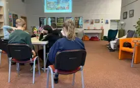 Uczestnicy podczas warsztat&oacute;w. Wszyscy siedzą na krzesłach, na ścianie wyświetlana prezentacja multimedialna.