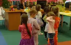 Dzieci stoją w kolejce do wypożyczenia książek. Bibliotekarka siedzi przy komputerze.