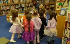 Dzieci oglądają książki ułożone w drewnianych kasetonach. Bibliotekarka tłumaczy, gdzie się znajdują poszczeg&oacute;lne bajki.