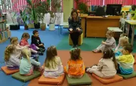 Dzieci siedzą w p&oacute;łkolu na dywanie na kolorowych poduszkach. Bibliotekarka czyta im książeczkę.