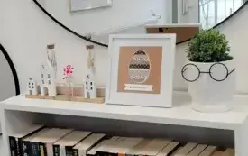 Tr&oacute;jwymiarowa kartka wielkanocna w białej ramce stoi na białej komodzie z książkami. Nad nią jest kawałek lustra, w kt&oacute;rym odbija się kawałek wnętrza biblioteki od strony wejścia.