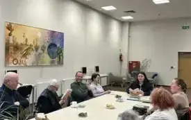 Przy długim stole w Wypożyczalni Gł&oacute;wnej siedzą uczestnicy Dyskusyjnego Klubu Książki. Przed nimi leżą egzemplarze omawianej książki oraz filiżanki. Po lewej stronie na ścianie wisi duży obraz.