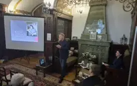 Zdjęcie wnętrza stylowego salonu . Po lewej stoi ekran projekcyjny z prezentacją; na slajdzie jest czarno-białe zdjęcie osoby oraz duży napis. Na środku kadru stoi mężczyzna w ciemnej marynarce i dżinsach, trzyma mikrofon i m&oacute;wi do zebranych. Wok&oacute;ł siedzi