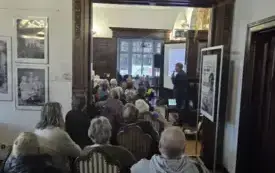  Pomieszczenie z publicznością siedzącą na krzesłach, fotografowana od tyłu przez duże, ozdobne drewniane przejście. W pomieszczeniu widać kilkanaście os&oacute;b, gł&oacute;wnie starszych, siedzących w kilku rzędach i zwr&oacute;conych w stronę prelegenta. Po prawej stronie 