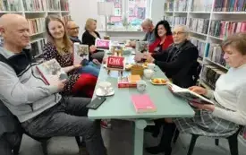 Przy stole siedzi ośmioro członk&oacute;w Dyskusyjnego Klubu Książki: czterech mężczyzn i cztery kobiety. Jedna z kobiet, kt&oacute;ra siedzi po prawej stronie, czyta książkę Magdaleny Grzebałkowskiej &bdquo;Dezorientacje. Biografia Marii Konopnickiej&rdquo;. Tr&oacute;jka z uczestnik