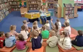 Dzieci siedzą na dywanie na wprost bibliotekarki, kt&oacute;ra pokazuje im obrazek i zadaje pytania, a dzieci podnoszą rączki do odpowiedzi.