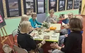 Klubowiczki siedzą dookoła stołu. Na stole jest jasnozielony obrus i poczęstunek.