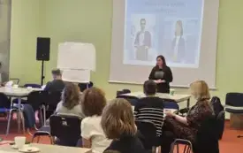 przy stolach siedzą uczestnicy spotkania, frontem do nich stoi kobieta i przemawia, za nią slajd z prezentacji wyświetlany na ścianie