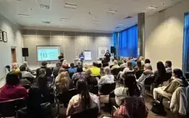 Na zdjęciu widać uczestnik&oacute;w spotkania, kt&oacute;rzy siedzą przodem do prowadzących warsztaty. Z przodu sali stoją prowadzące (dr Justyna Bucknall-Hołyńska, Anna Bezdziecka) oraz tłumaczkę PJM Joannę Grochalę,  widać też za ich plecami przy ścianie ekran, a po 