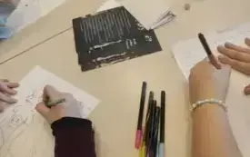 na stoliku leżą przybory do pisania i rysowania, wok&oacute;ł blat&oacute;w widać ręce uczestnik&oacute;w, na blacie zaś widać karki z ilustracjami do legend