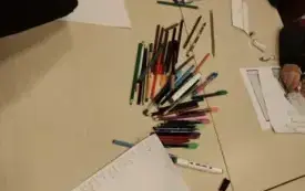 na stoliku leżą przybory do pisania i rysowania, wok&oacute;ł blat&oacute;w widać ręce uczestnik&oacute;w, na blacie zaś widać karki z tekstem i ilustracjami do legend