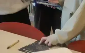  uczestniczka trzyma w jednej dłoni telefon z grą i wskaz&oacute;wkami, drugą rękę opiera na granatowej kartce z instrukcjami. Przed nią na stoliku odwijająca się z rulonu kartka a4