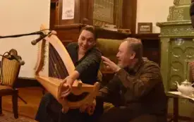 Kobieta i mężczyzna rozmawiają, kobieta trzyma harfę, w tle kominek