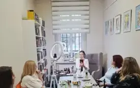Zdjęcie przedstawia kameralne spotkanie. Kobieta w białym stroju siezi w szarym fotelu i wykonuje makijaż na swojej twarzy, demonstrując techniki grupie