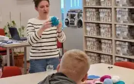 Przy stole stoi bibliotekarka i pkazuje maskotkę z wł&oacute;czki, przy stole siedzą uczniowie