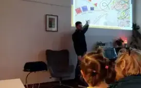 Prowadzący wskazuje dłonią na ścianie, na kt&oacute;rej wyświetlona jest prezentacja z kolorowymi elementami.