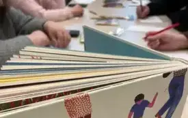 Na stole stoi książka, rozłożona w formie harmonijki, w tle widoczny długi st&oacute;ł, kredki i ręce uczestnik&oacute;w spotkania.
