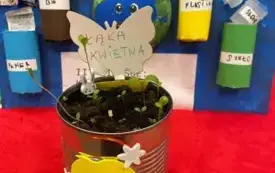 Na pierwszym planie puszka ziemia, kiełkujące roślinki, drewniany motylek, etykieta z napisem łąka kwietna. W tle praca z przyklejonymi kolorowymi rolkami, z kt&oacute;rych wystają odpowiednio posegregowane śmieci.