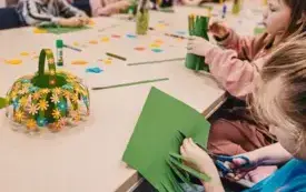 Dzieci siedzą przy stołach i tworzą z kolorowego papieru koszyczki z kwiatami. 