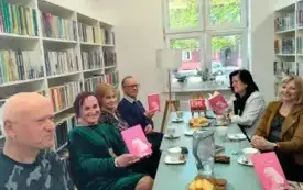 Na zdjęciu widać sz&oacute;stkę klubowicz&oacute;w Dyskusyjnego Klubu Książki: czworo z lewej i dwie klubowiczki z prawej. Jedna z klubowiczek czyta książkę. Na stole jest mała sztaluga z logo Dyskusyjnego Klubu Książki, słodycze, filiżanki, talerzyki, książki, dzba