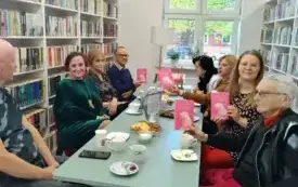 Przy stole siedzi ośmioro członk&oacute;w Dyskusyjnego Klubu Książki: trzech mężczyzn i pięć kobiet. Cztery osoby z lewej strony i cztery osoby z prawej strony. Czw&oacute;rka klubowicz&oacute;w trzyma w g&oacute;rze książki Nataszy Sochy &bdquo;Udupione przez matki&rdquo;. Na stole jest mał