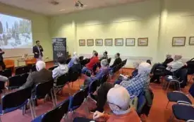 Zdjęcie zrobione od boku przedstawia całą salę z uczestnikami spotkania. Po lewej stronie prelegent i moderator. W środku uczestnicy siedzący na krzesłach. Wśr&oacute;d nich po prawej stronie widać dyrektora biblioteki.