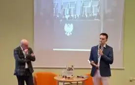Na środku zdjęcia stoją prelegent oraz moderator spotkania. Obok nich stolik i dwa fotele. Za nimi wyświetlona jest prezentacja prelegenta &ndash; widać na niej zdjęcie r&oacute;żnych postaci. Moderator m&oacute;wi do mikrofonu.