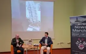 Na środku zdjęcia, na fotelach przy stoliku siedzą prelegent oraz moderator spotkania. Prelegent m&oacute;wi do mikrofonu, a na stoliku przy nim prezentuje się książka jego autorstwa. W tle wyświetlona jest prezentacja. Po prawej stronie stoi rollup z nazwą cykl