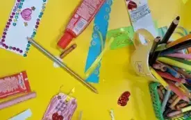 Dwoje dzieci ozdabia swoje zakładki do książek. Na stole leżą nożyczki, kleje, wycinanki oraz kolorowe kredki.