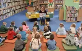 Bibliotekarka omawia z dziećmi historię pisarza, pokazując obrazek w książce. Dzieci siedzą na dywanie na kolorowych poduchach. W tle znajdują się regały z książkami.
