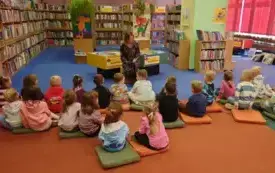 Dzieci siedzą na kolorowych poduchach na dywanie przed bibliotekarką i słuchają bajeczki. W tle znajdują się regały z książkami.