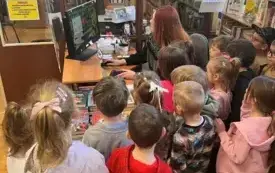 Kobieta przy biurku pokazuje zgromadzonym dzieciom program na komputerze