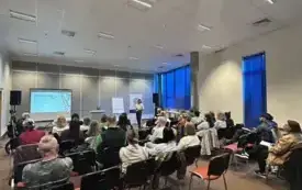 Na zdjęciu widać uczestnik&oacute;w spotkania, kt&oacute;rzy siedzą przodem do prowadzącej warsztaty. Z przodu sali stoi prowadząca dr Justyna Bucknall-Hołyńska. Widać też po lewej stronie ekran, a po prawej okna.