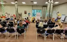 Dzieci i rodzice siedzą na krzesłach i słuchają opowieści autorki podczas spotkania w Oddziale Dziecięcym.