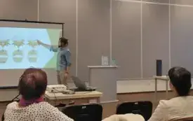 Na zdjęciu na pierwszym planie widzimy pomarańczowe krzesła, na kt&oacute;rych siedzą uczestnicy spotkania. W tle prowadzący pokazuje na rzutniku rysunki kręg&oacute;w kręgosłupa. Obok stoi podest z książką i dwiema butelkami wody.
