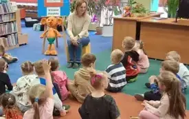 Dzieci siedzą na dywanie i zgłaszają się do odpowiedzi podczas pogadanki. Obok bibliotekarki na sztaludze stoi antyrama z plakatem XXIII Og&oacute;lnopolskiego Tygodnia Bibliotek i z nazwą zajęć.