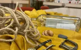 Na udrapowanym obrusie leżą rekwizyty: guzik, stary damski zegarek, klamra od paska męskiego, butelka z listem w środku, muszla, spinka do włos&oacute;w z zielonymi kamieniami.