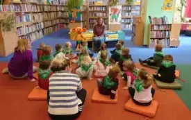 Bibliotekarka prowadzi pogadankę, obok niej stoi Plastuś. Dzieci siedzą na podłodze na kolorowych poduchach. W tle znajdują się regały z książkami.