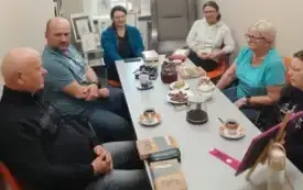 Spotkanie Dyskusyjnego Klubu Książki w bibliotece. Pięć uczestniczek siedzi wok&oacute;ł podłużnego stołu, na kt&oacute;rym leżą omawiane książki, filiżanki i dzbanki z herbatą. Kobiety rozmawiają w przyjaznej atmosferze. W tle, na sztaludze, stoi tabliczka z r&oacute;żowym l