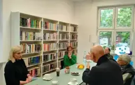 Sz&oacute;stka uczestnik&oacute;w spotkania siedzi przy stole i dyskutuje. Na stole są białe filiżanki, talerzyki, butelka z wodą, słodycze i skoroszyt. Po lewej stronie widać cztery białe regały z książkami.  Z tyłu na środku widać klosz białej lampy i okno Jedna z