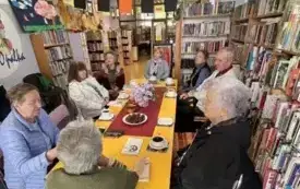 Siedem kobiet i mężczyzna siedzą przy stole, na nim bukiet bzu, książki,słodycze i filiżanki