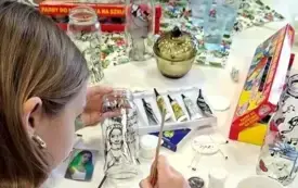 Dziewczyna maluje szklaną butelkę. Na stole obok niej znajdują się tubki i słoiczki z farbami oraz filiżanka z herbatą, a w oddali &ndash; wazon z bzem.