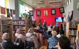 Grupa os&oacute;b w bibliotece, trwa spotkanie. W tle kobieta stoi i opowiada o wyświetlanych zdjęciach