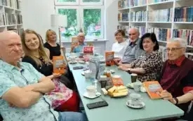 Dziewiątka uczestnik&oacute;w spotkania Dyskusyjnego Klubu Książki (pięć kobiet, czw&oacute;rka mężczyzn) siedzi przy miętowym stole. Czw&oacute;rka klubowicz&oacute;w trzyma w g&oacute;rze książkę &bdquo;Dokt&oacute;rka od familok&oacute;w&rdquo; Magdaleny Majcher. Na stole są białe filiżanki, talerzyki, słodyc