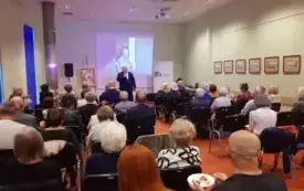 Publiczność siedzi na krzesłach i słucha dyrektora Sławomira Szenwalda przemawiającego do mikrofonu.
