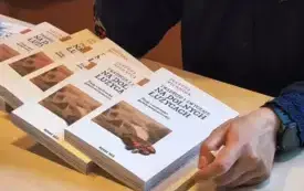 Zdjęcie przedstawia zbliżenie na st&oacute;ł. Leżą na nim egzemplarze książki wydanej przez prelegentkę. Autorka siedzi przy stole, widać jej dłoń trzymającą długopis i fragment ozdobnej koszuli.