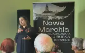Zdjęcie przedstawia zbliżenie na prelegentkę. W jednej ręce trzyma mikrofon, a drugą gestykuluje. Obok niej stoi rollup z nazwą cyklu spotkań.