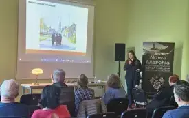 Na pierwszym planie znajdują się krzesełka, na kt&oacute;rych siedzą uczestnicy spotkania. Ponad nimi widać prezentację ze zdjęciem przedstawiającym jeden z wiosennych zwyczaj&oacute;w Serbołużyczan. Po prawej stronie stoi prelegentka oraz rollup z nazwą cyklu spotkań.