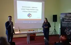 Na pierwszym planie, patrząc od lewej strony, znajdują się kierownik Działu Zbior&oacute;w regionalnych rozpoczynający spotkanie oraz prelegentka. Za nimi jest st&oacute;ł z wyeksponowaną książką autorstwa prelegentki. Dalej na ścianie wyświetlona jest tytułowa strona 